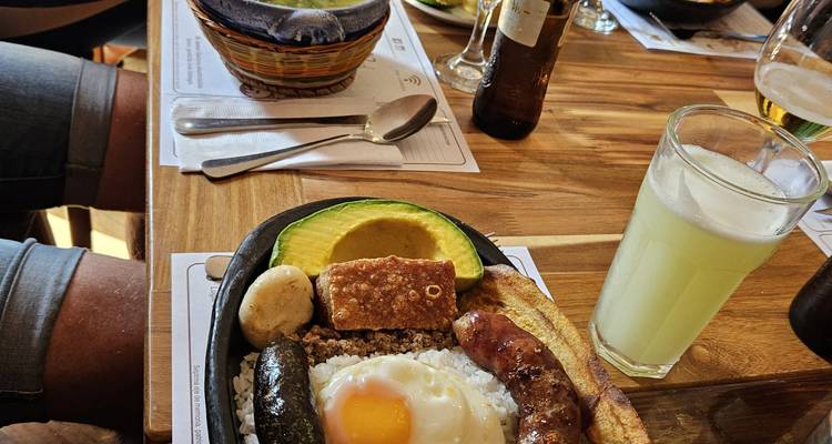 Gros plan d'un copieux repas colombien bandeja paisa avec avocat, œuf frit, banane plantain et saucisse servi sur une table en bois aux côtés d'un jus frais et d'une bière.