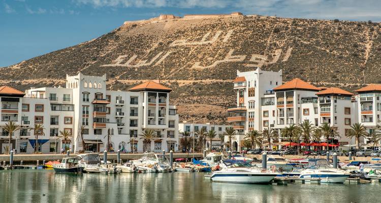Moderner Jachthafen mit weißen Apartments im mediterranen Stil unterhalb eines Hangs mit riesiger arabischer Schrift in Agadir.