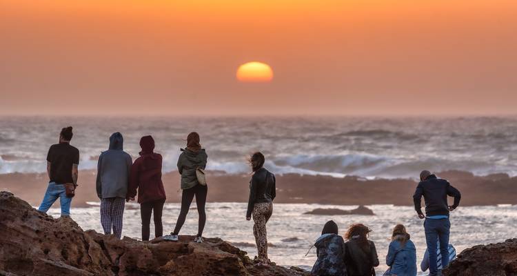 Silhouetten von Menschen, die auf felsigem Ufer stehen und einen orangen Sonnenuntergang über dem Atlantischen Ozean beobachten.