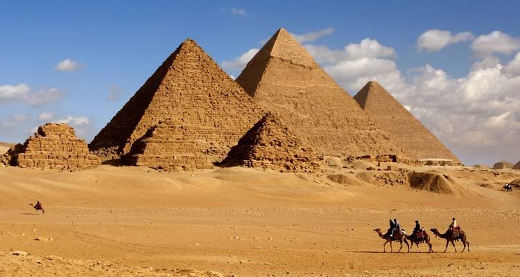 Camel caravan traversing the sandy foreground with the three great pyramids towering behind
