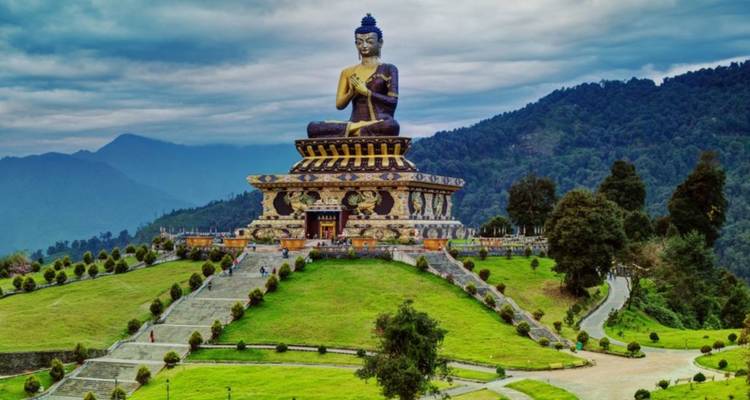 Large seated Buddha statue atop an ornate pedestal surrounded by manicured lawns and forested hills