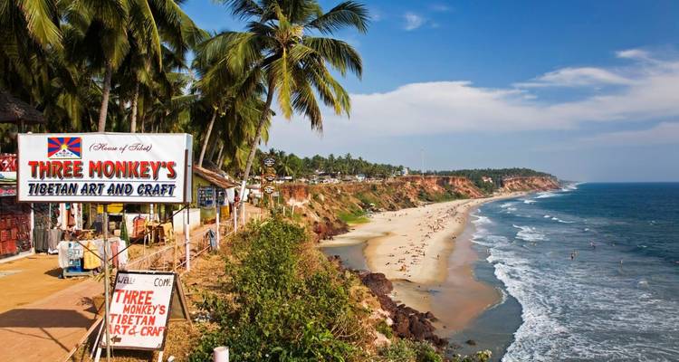 Cliff-top coastal town with palm trees and beach below, viewed past colorful roadside craft shops.