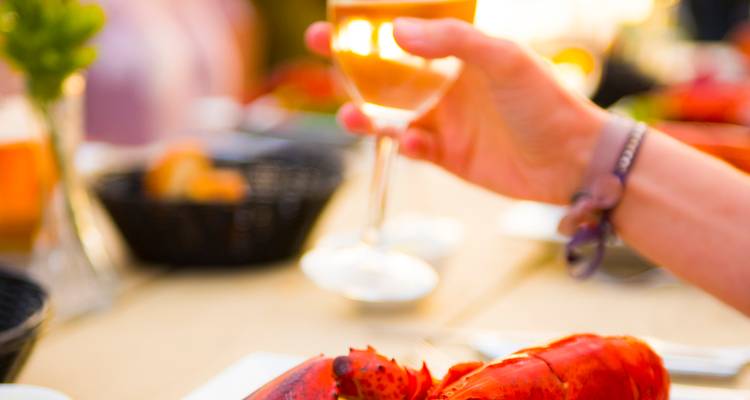 Close-up of a diner toasting white wine beside a freshly cooked lobster meal.