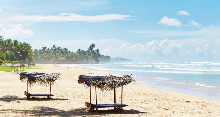 Ruhiger tropischer Strand mit zwei einfachen strohgedeckten Sonnenschutzunterständen vor sanfter Brandung unter blauem Himmel.