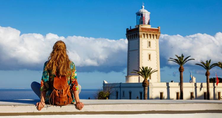 Traveler with backpack sits on a white wall overlooking Cap Spartel lighthouse, palm trees and distant sea