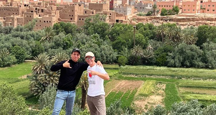 Two friends pose with thumbs up overlooking a lush oasis and adobe town