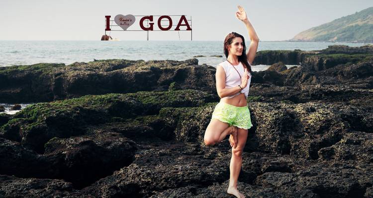 Young woman practices a yoga pose on rugged coastal rocks with an 'I love Goa' sign above the sea.