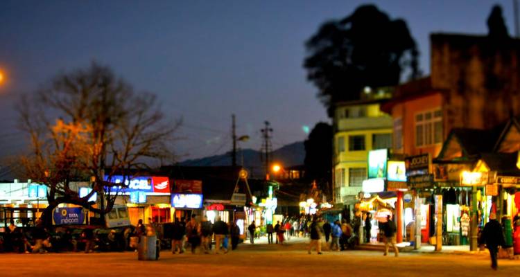 A lively hill-town street glows with shop lights and evening crowds against a twilight sky.