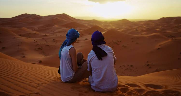 Two travellers in turbans sit on a high dune overlooking endless desert ridges at sunset.