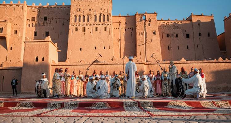 Traditional musicians and dancers perform in colourful robes before a grand earthen kasbah under blue skies.
