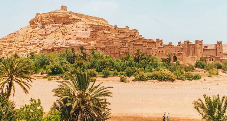 A fortified clay village crowns a hill above palm groves and a dry riverbed.