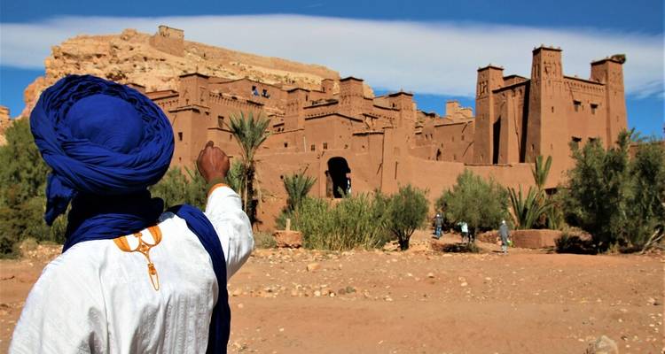 A Berber guide in blue scarf gestures toward an ancient earthen kasbah complex.
