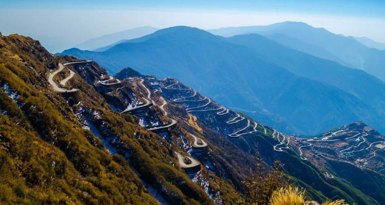 A dramatic zig-zag mountain road snakes along steep slopes with hazy blue ranges beyond.