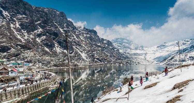 Tourists play in snow beside a reflective alpine lake framed by rugged peaks.