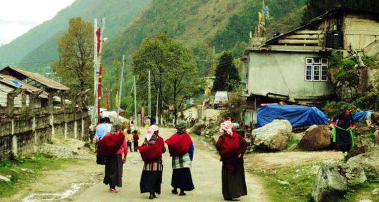 Local women in red shawls walk along a rural mountain road past simple houses.