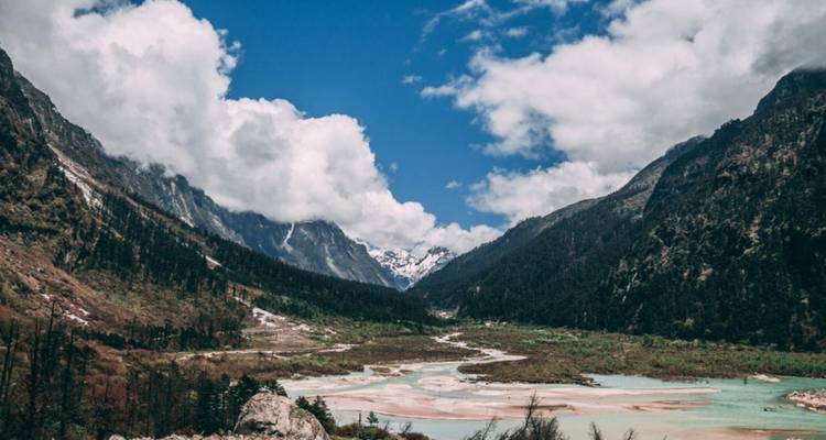 Wide glacial valley with turquoise braided river beneath towering peaks and clouds.