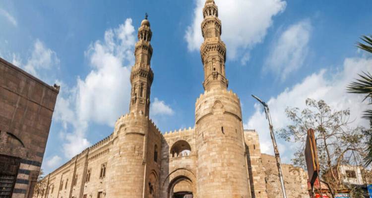 Stone mosque with two ornate minarets set against a bright blue sky with scattered clouds.