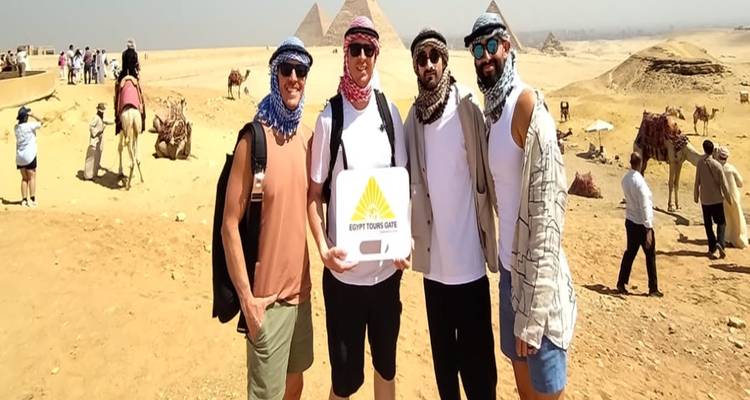 Four young men in desert scarves posing with a tour sign in front of the distant pyramids and camels.