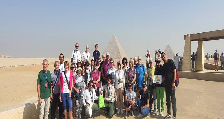 Group of tourists kneeling and standing together for a photo with pyramids in the distance.