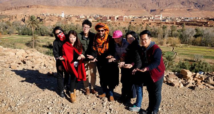 Travelers in bright scarves pose with open palms overlooking a desert village and oasis.