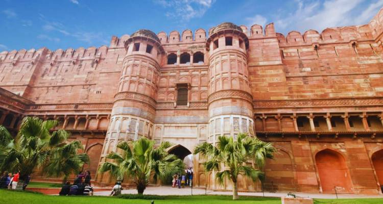 Massive red-sandstone gateway and twin bastions of a fort with tourists milling on lush lawns below a blue sky.