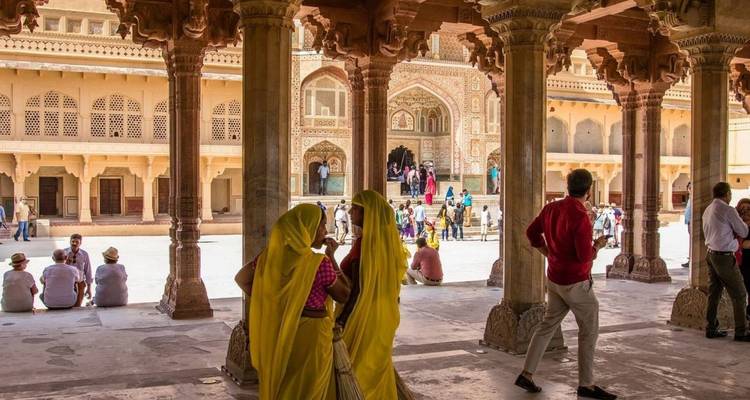 Courtyard of a palace with carved columns framing visitors; two women in bright yellow saris converse in foreground.