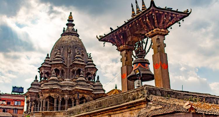 Stone bell pavilion and ornate temple spire adorned with small sculptures beneath a partly cloudy sky.