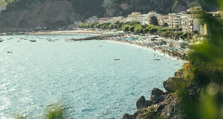 Plage animée et station balnéaire entourée de collines boisées, petits bateaux sur une eau scintillante.