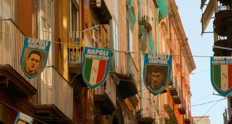 Rue étroite ornée de bannières du championnat de football de Napoli bleu entre des balcons colorés.