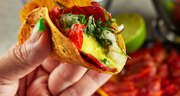 Hand mit einem frisch zubereiteten Taco al Pastor, garniert mit Avocado, Zwiebel und Koriander.