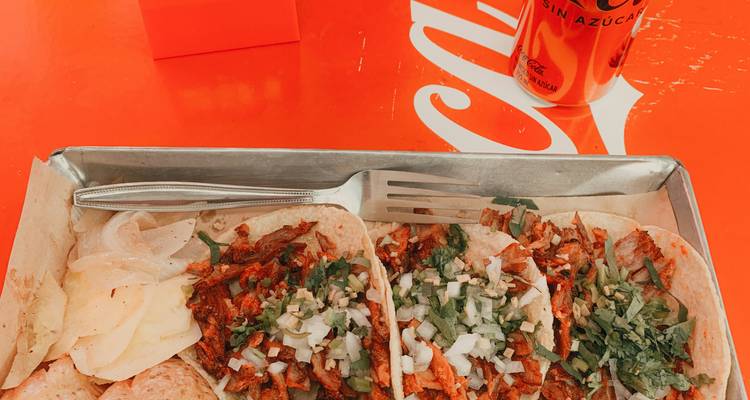 Tablett mit Tacos, serviert mit gehackter Zwiebel und Koriander auf einem leuchtend orangefarbenen Tisch neben einer Cola.