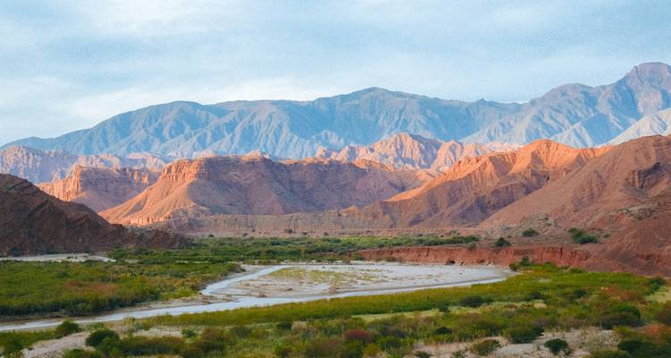 Wide Andean valley with red rock formations, green riverbed and distant blue mountains.