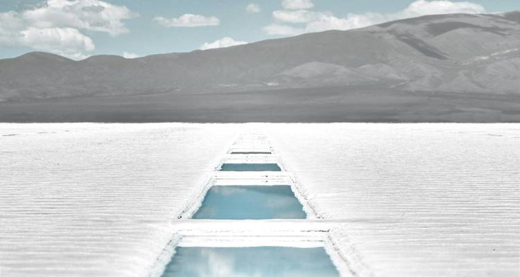 Straight salt-flat channel filled with blue water stretching toward distant Andean mountains under bright sky.