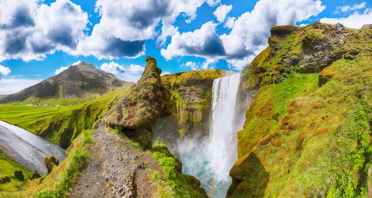 Wide panoramic view of Skogafoss waterfall plunging over green cliffs under a bright blue sky.