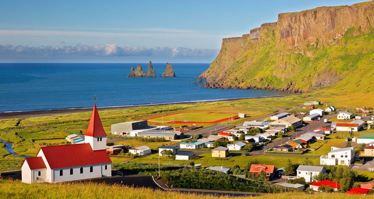 Vue panoramique sur l'église de Vik en direction de la mer et des falaises de Reynisdrangar par une matinée dégagée.