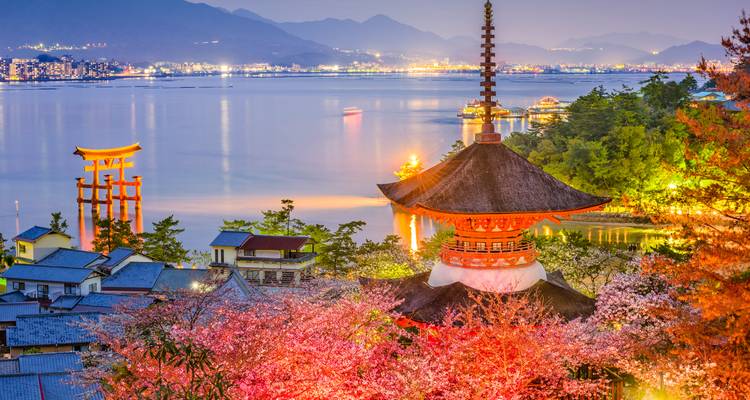Twilight panorama of Miyajima with five-story pagoda, illuminated torii gate and cherry blossoms overlooking the bay.