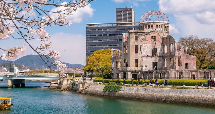The Atomic Bomb Dome stands beside a tranquil river framed by cherry blossoms and city skyline in Hiroshima.
