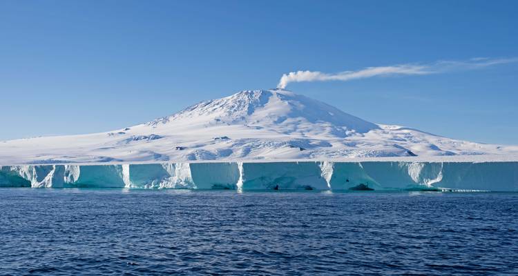 Snow-covered Mount Erebus emitting a small plume above a sheer ice shelf rising from a calm blue sea under clear sky.