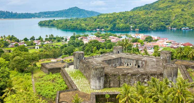 Aerial view of massive stone fort overlooking a lush volcanic island, colorful village and tranquil bay.