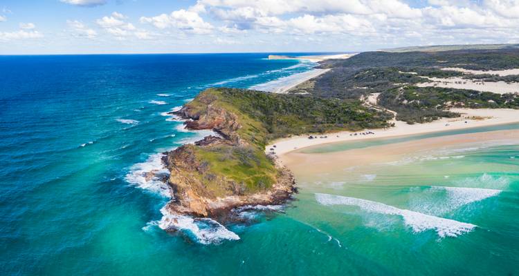 Aerial view of rugged green headland and turquoise waters along a pristine sandy coast.