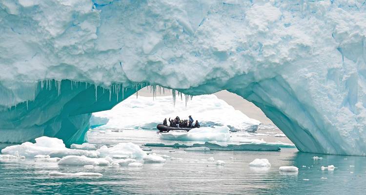 Zodiac boat with bundled passengers passes under a dramatic turquoise ice arch amid Antarctic ice floes.