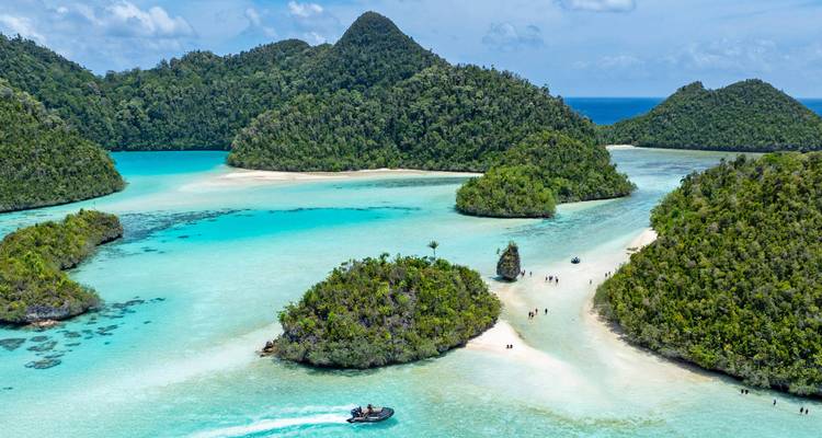 Crystal-clear turquoise channels winding between lush green limestone islets with a small speedboat and beachgoers, Raja Ampat.