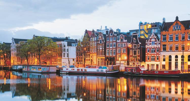 Row of illuminated canal houses and boats mirrored in calm Amsterdam waters at dusk.