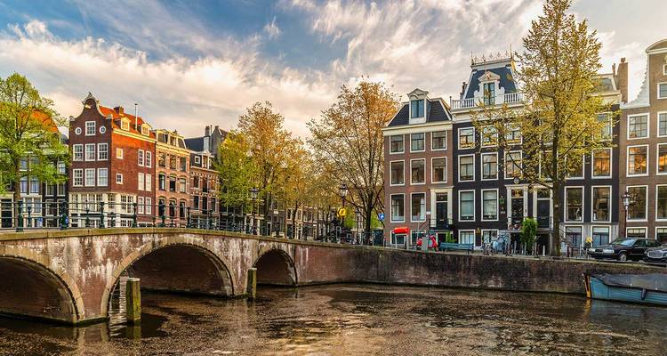 Picturesque Amsterdam canal scene with arched brick bridge, historic gabled houses and warm evening light.
