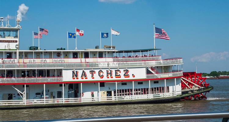 El histórico barco de vapor 'Natchez' navegando por el río Mississippi bajo un cielo azul despejado con múltiples banderas ondeando.