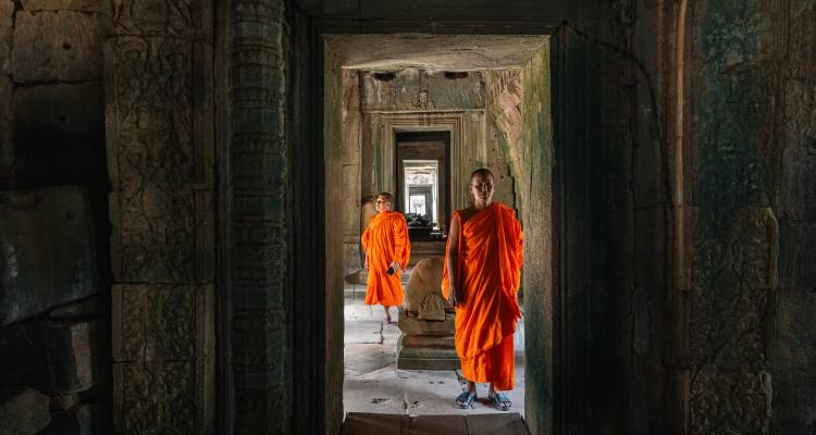 Two Buddhist monks in bright orange robes standing inside a dim stone corridor of Angkor Wat.