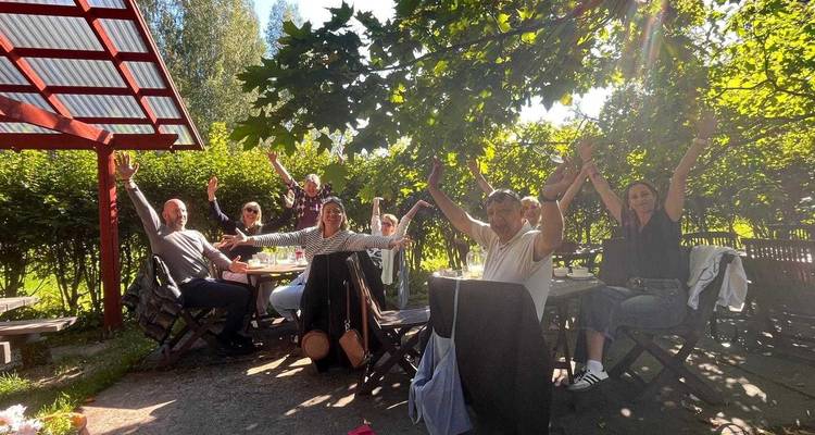 A cheerful group raises their arms around an outdoor garden table enjoying food and sunshine.