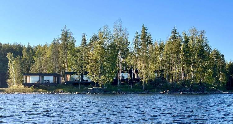 Modern black cabins nestle among tall pine trees on a small island surrounded by blue lake waters.