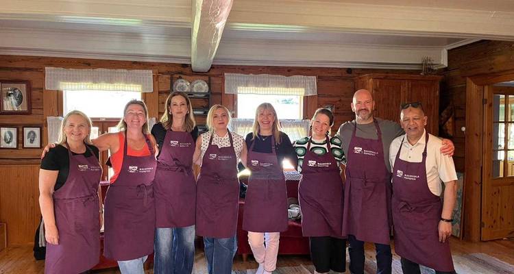 Participants in matching aprons pose inside a rustic wooden house before a cooking class.