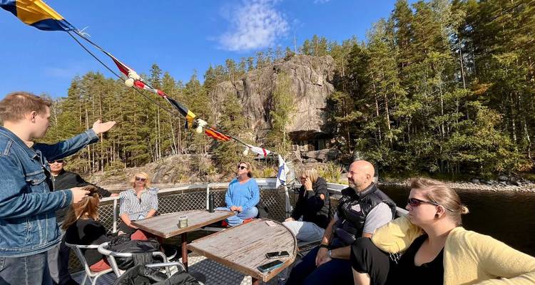 Tourists relax at tables on an open boat deck cruising past forested cliffs on a sunny day.